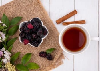 Vista superior de uma xícara de chá escura ao lado de uma tigela branca em formato de flor cheia de amoras pretas e vermelhas frescas, decorada com paus de canela e flores delicadas sobre uma mesa branca.