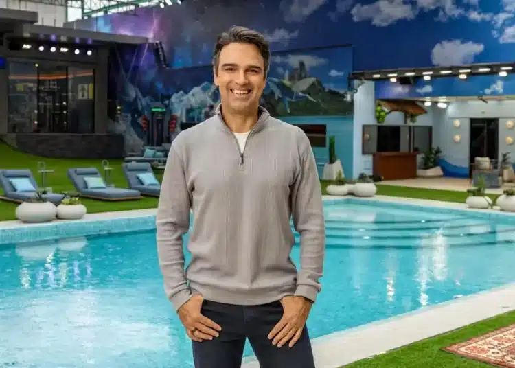Tadeu Schmidt sorrindo na área externa da casa do BBB 26, com piscina ao fundo.