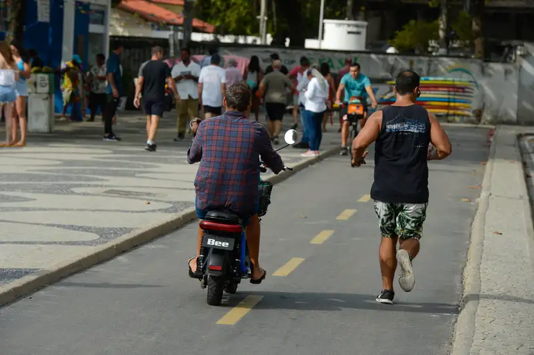 Pessoa em ciclomotor no meio de ciclistas e pedestres, destacando a necessidade de registro, placa e CNH, conforme as novas regras.