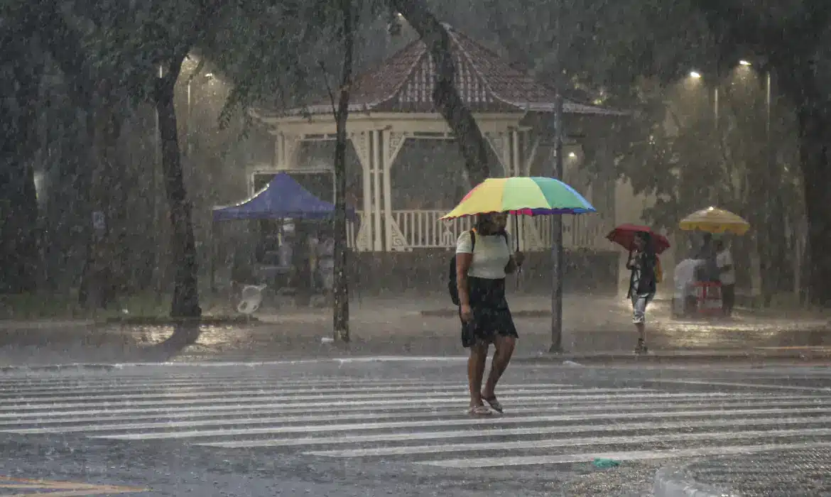 Mulher atravessa rua com guarda-chuva colorido durante intensa chuva em área urbana