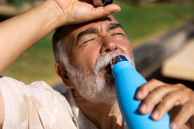 Homem idoso com barba branca bebendo água de uma garrafa térmica azul, com os olhos fechados e a mão na testa, aliviando-se do calor em um ambiente externo.
