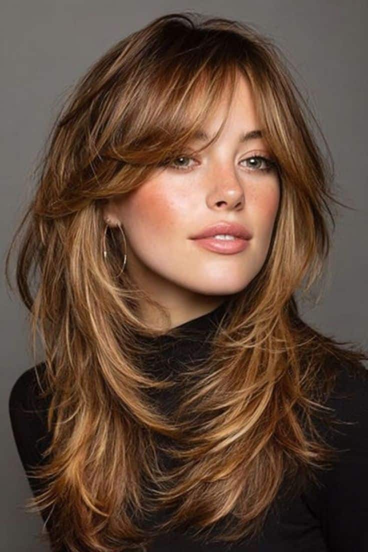 Mulher de cabelos castanhos claros com luzes mel, exibindo o corte Butterfly Cut com camadas longas e franja cortina que emoldura o rosto.
