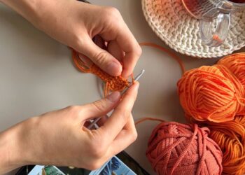 Mãos fazendo crochê com fios coloridos, tablet com inspirações e chá ao lado