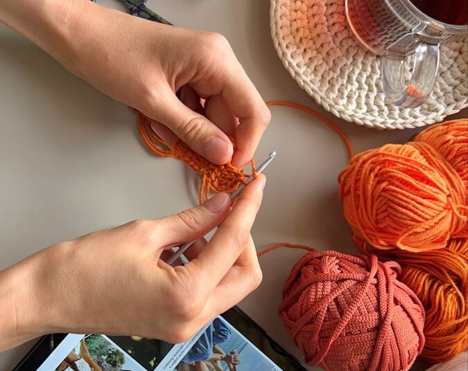 Mãos fazendo crochê com fios coloridos, tablet com inspirações e chá ao lado