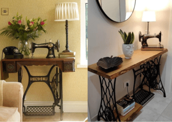 Máquina de costura Singer antiga de pedal com a base de ferro trabalhada e um tampo de madeira, transformada em um aparador com vaso de flores, luminária e telefone retrô.