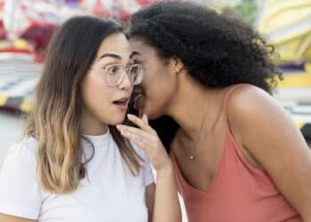 Duas mulheres jovens em um parque de diversões, uma delas sussurrando algo no ouvido da outra, que reage com expressão de surpresa.