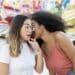Duas mulheres jovens em um parque de diversões, uma delas sussurrando algo no ouvido da outra, que reage com expressão de surpresa.