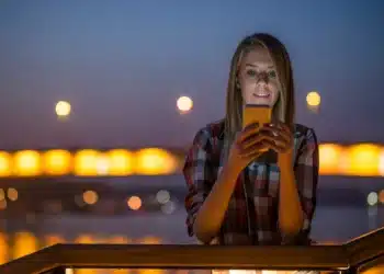 Mulher sorrindo e usando celular à noite, com luzes suaves ao fundo, ideal para compartilhar mensagens inspiradoras.