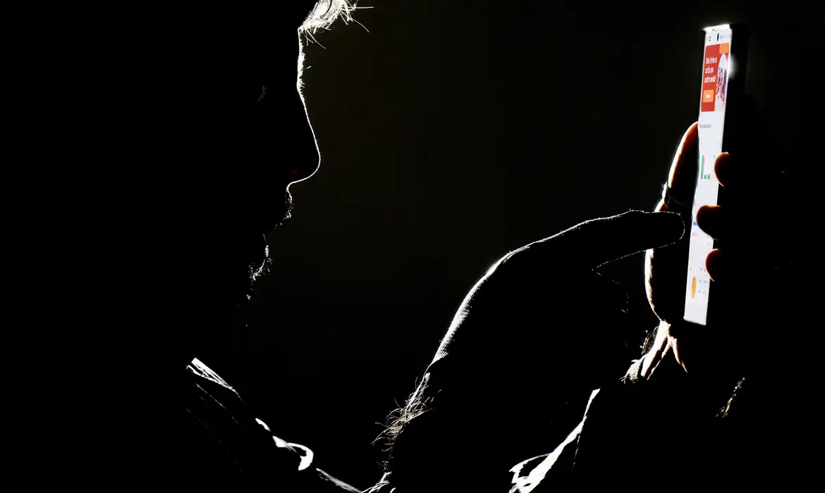 Silhueta de homem mexendo no celular iluminado em fundo preto