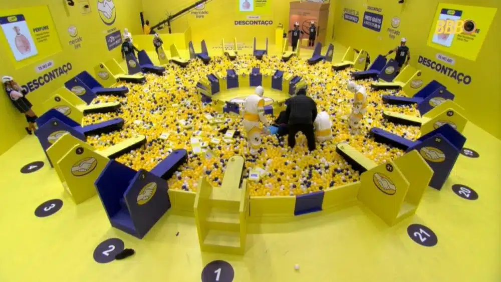 Vista aérea do cenário circular da prova de resistência do BBB 26, com temática amarela do patrocinador Mercado Livre. No centro da piscina de bolinhas amarela e branca, participantes em uniformes brancos e pretos e a equipe médica prestam socorro a Henri Castelli, que está deitado após sofrer uma convulsão.