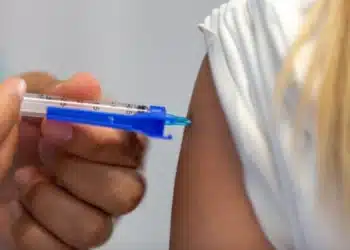 Mão aplicando vacina com seringa azul no braço de uma pessoa com camiseta branca