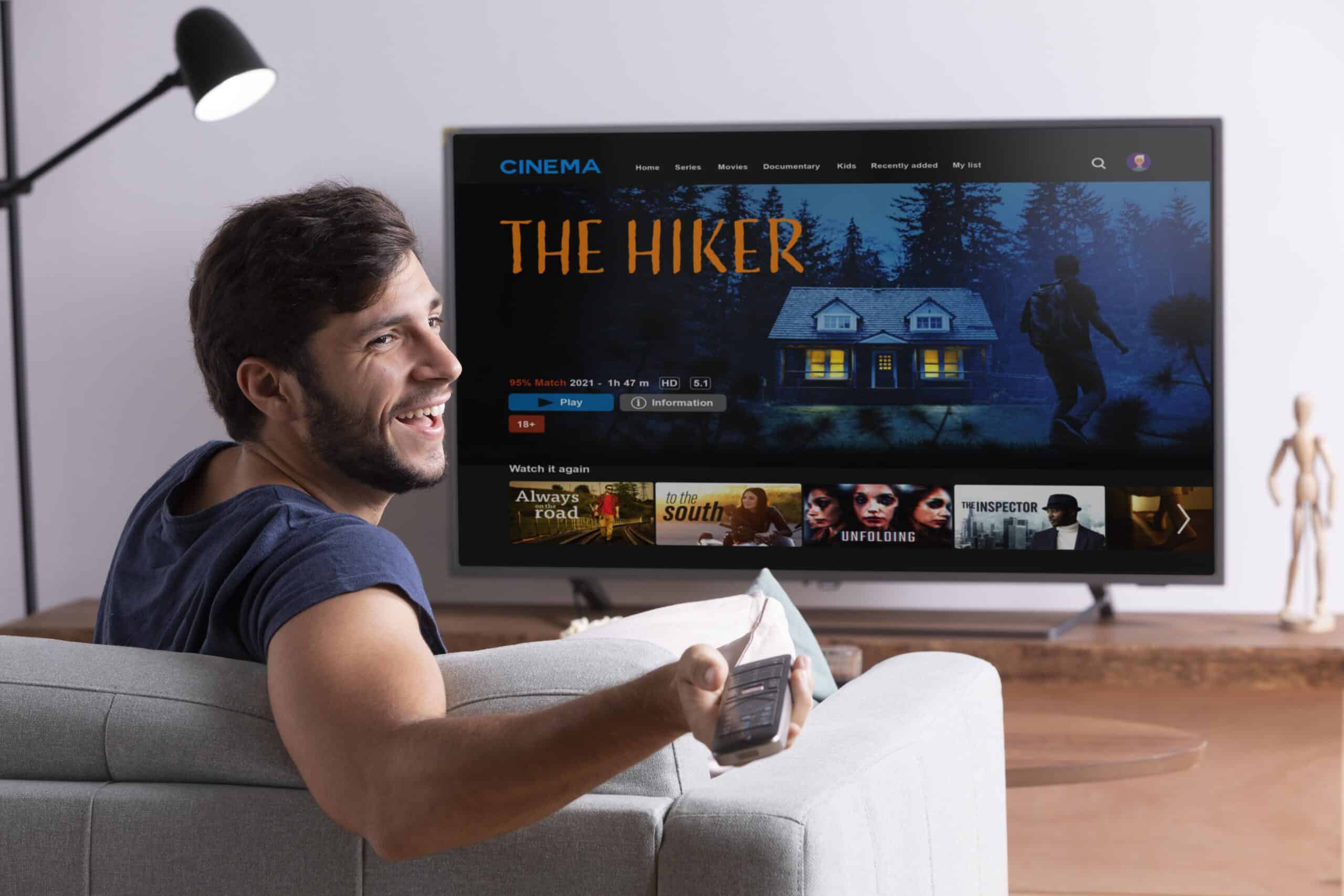 Homem sorridente no sofá com controle remoto assistindo o filme The Hiker na televisão