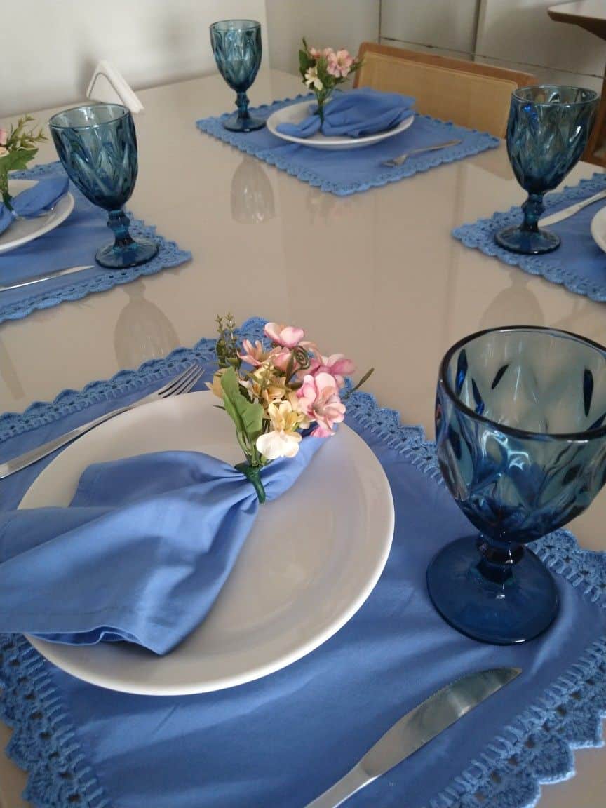 Mesa posta com sousplats azuis, taças azul escuro, guardanapos e flores.