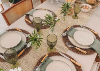 Mesa de jantar decorada com sousplats de rattan, louças claras, taças verdes e detalhes de plantas naturais