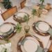 Mesa de jantar decorada com sousplats de rattan, louças claras, taças verdes e detalhes de plantas naturais