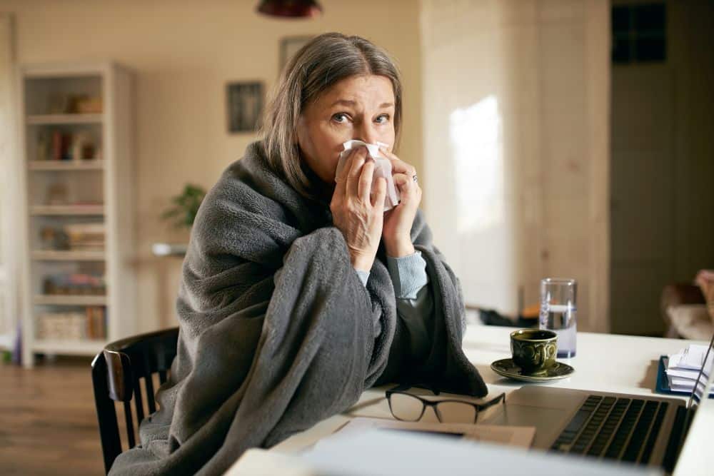 Mulher idosa sentada à mesa, envolta em um cobertor cinza, usando um lenço para cobrir o nariz e a boca; à sua frente estão um notebook, uma xícara de café e óculos de grau.