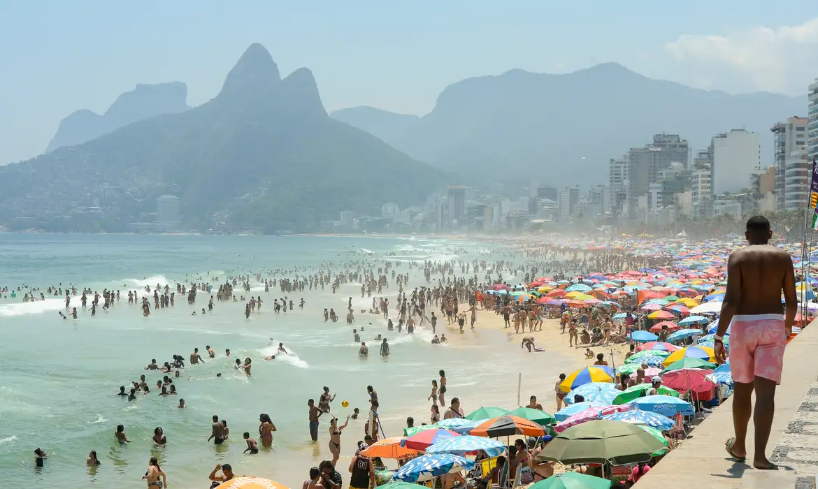 Praia de Ipanema cheia de banhistas e guarda-sóis coloridos com montanhas Dois Irmãos ao fundo