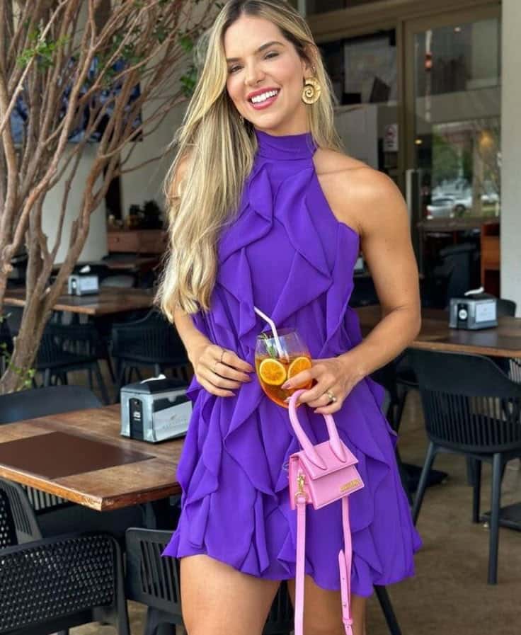 Mulher loira sorridente vestindo um vestido curto roxo vibrante com babados verticais e gola alta. Ela segura uma mini bolsa rosa chiclete e um drink com rodelas de laranja, em um ambiente externo de restaurante.