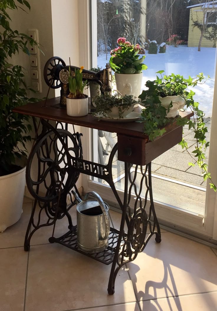 Base de ferro de uma máquina de costura Singer antiga, com tampo de madeira rústica, utilizada como suporte para vasos de plantas variadas em uma varanda ou ambiente com luz natural.