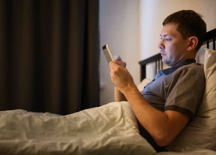 Homem deitado na cama à noite, em um quarto escuro, segurando um tablet iluminando seu rosto. Ele demonstra expressão séria enquanto usa o dispositivo antes de dormir.