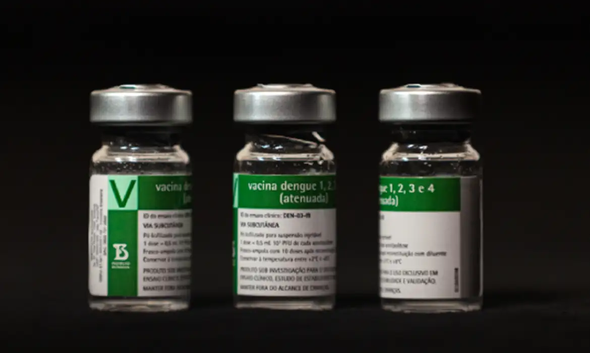 Três frascos transparentes da vacina contra dengue com rótulos verdes sobre fundo preto.