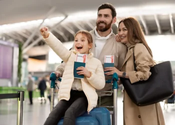 Família sorridente no aeroporto, com pai, mãe e filha segurando passagens de avião e com a criança empolgada sobre um carrinho de bagagem.