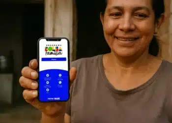Mulher sorridente mostrando smartphone com aplicativo Bolsa Família em frente à casa