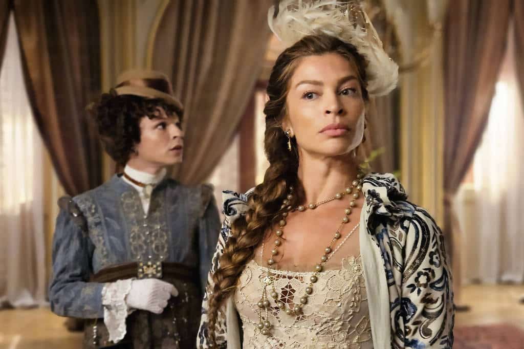 Duas mulheres usando roupas de época ricamente detalhadas em salão luxuoso