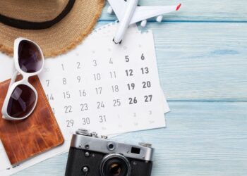 Chapéu, óculos escuros, passaporte, câmera e calendário sobre mesa de madeira.