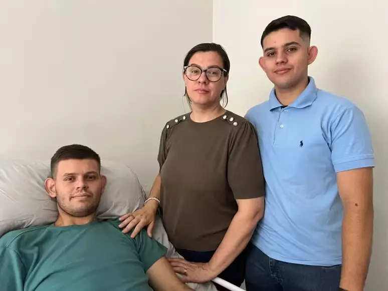 Mulher e jovem abraçam homem deitado em cama, expressando apoio e união no quarto.