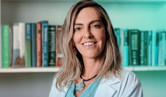 Mulher de jaleco branco sorri em frente a estante repleta de livros coloridos.