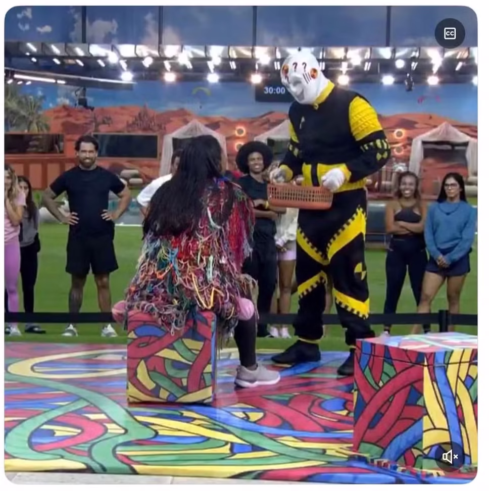  Jovem sentado diante de dummy do BBB segura mãos no peito e sorri com emoção