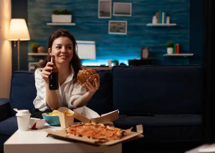 Mulher sorrindo e segurando uma garrafa de cerveja enquanto come fast food, com pizza e batatas fritas em uma mesa.