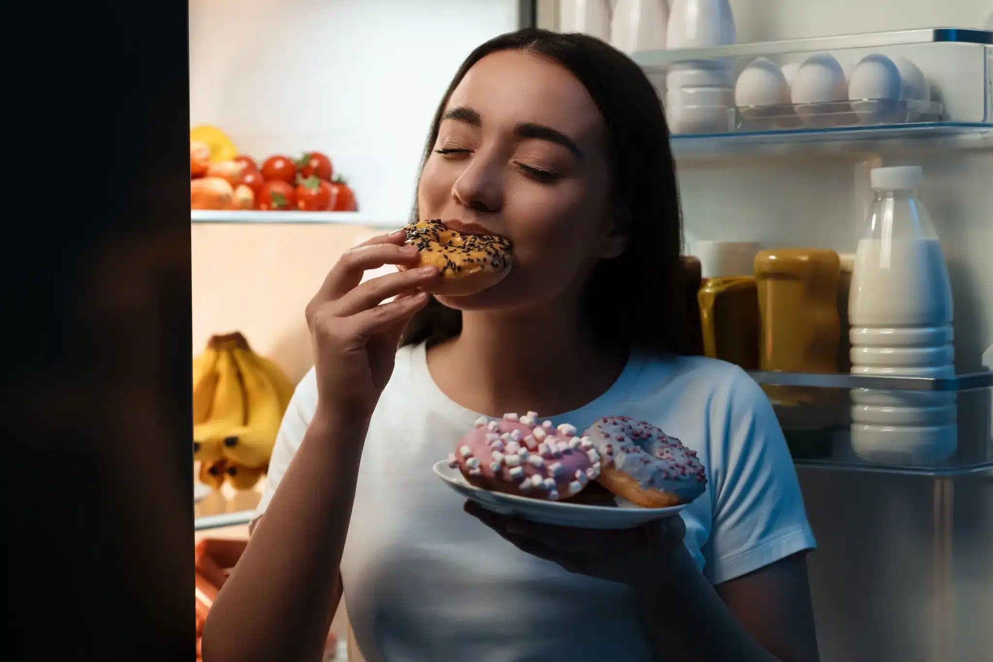 Mulher comendo donuts à noite em frente à geladeira, com expressão satisfeita.