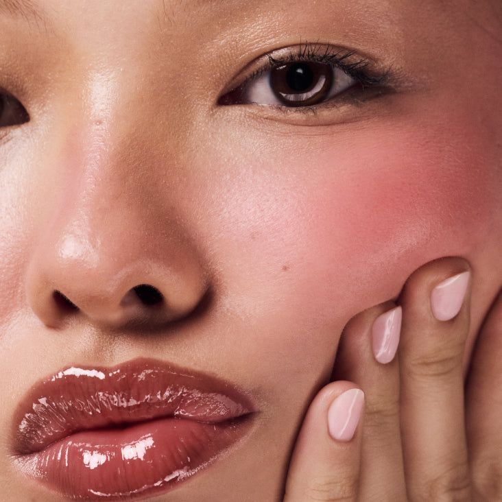 Rosto de mulher com pele iluminada, blush rosado e lábios gloss, mão tocando o rosto.