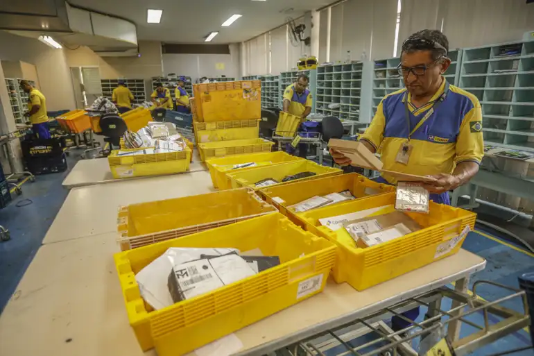 Funcionários dos Correios organizando entregas antes da volta das agências Funcionário dos Correios separa encomendas em caixas amarelas numa central de triagem