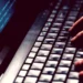 Alerta de golpe: criminosos exigem depósitos antecipados para liberar falsos valores judiciais. Imagem mostra mãos digitando em um teclado de notebook, enquanto a tela exibe códigos de programação iluminados, reforçando o caráter digital do golpe.