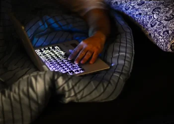 Pessoa usa notebook iluminado enquanto deita na cama com cobertores escuros, enquanto envia mensagens de boa noite para pessoas especiais