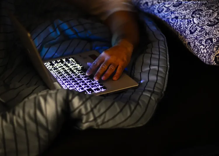 Pessoa usa notebook iluminado enquanto deita na cama com cobertores escuros, enquanto envia mensagens de boa noite para pessoas especiais