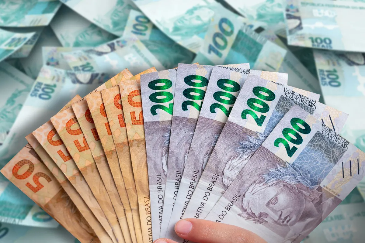 Mão exibe leque de notas de R$ 50 e R$ 200 sobre pilhas de dinheiro, após saque do microcrédito CadÚnico