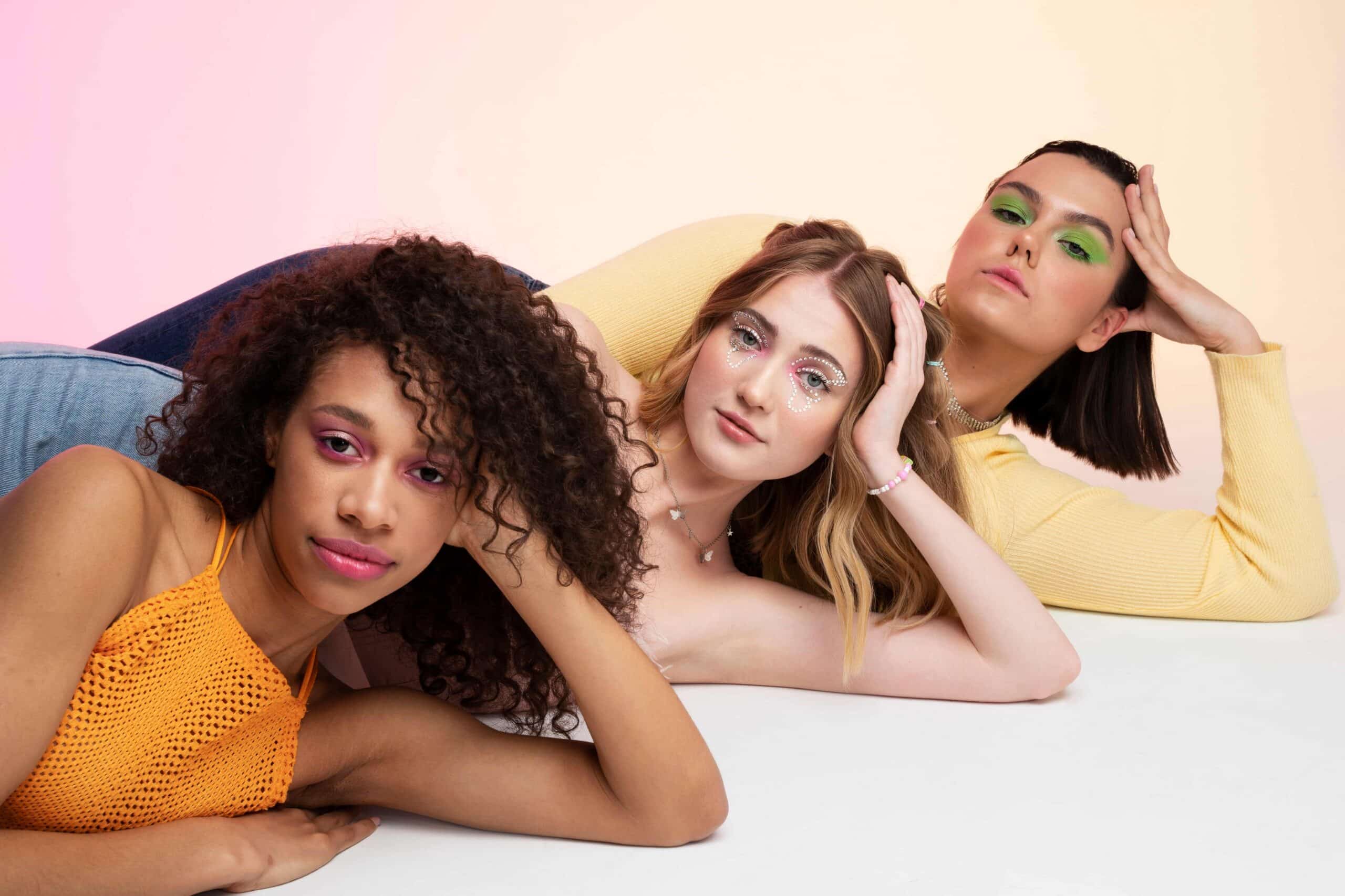 Três mulheres deitadas lado a lado, com maquiagem vibrante e poses marcantes