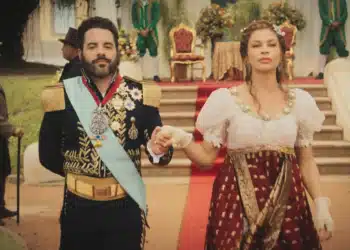 Homem com uniforme imperial caminha de mãos dadas com mulher em vestido de época diante de trono e tapete vermelho.