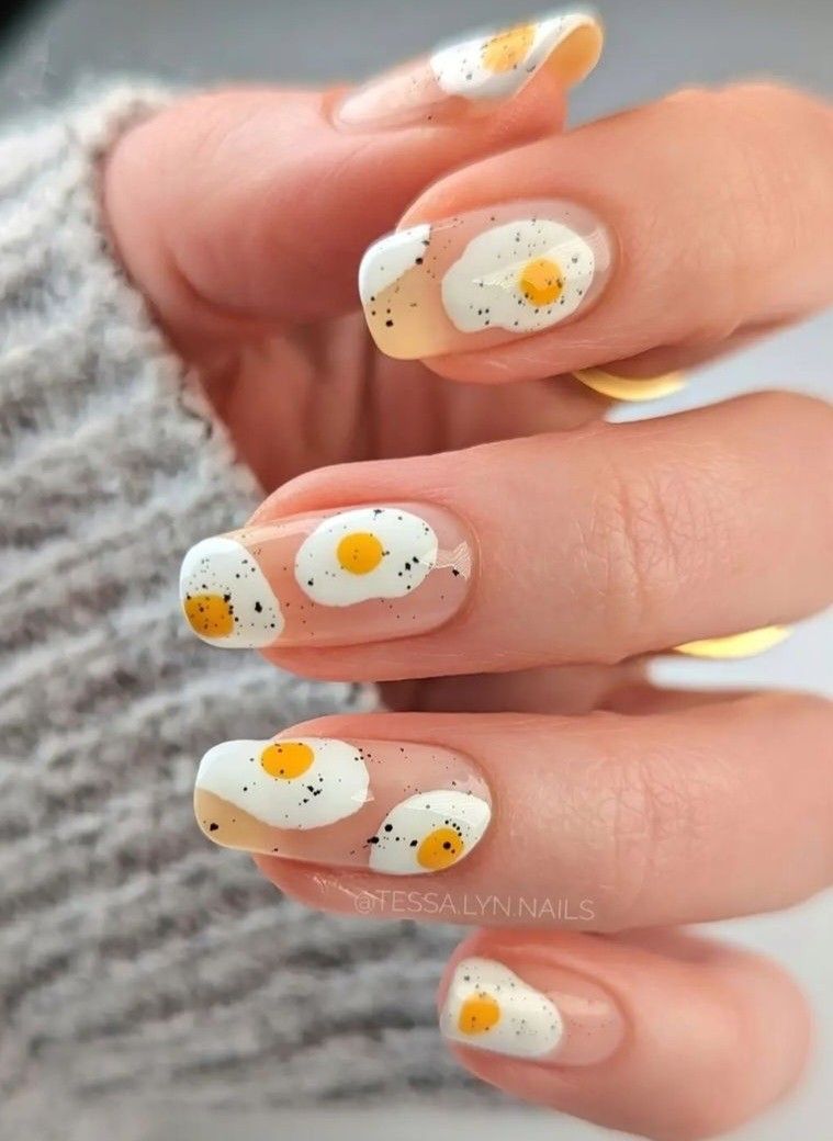 Mão com unhas pintadas de desenhos de ovos fritos, com detalhes em branco, amarelo e preto.