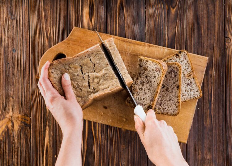 Mãos cortando pão integral em fatias sobre tábua de madeira