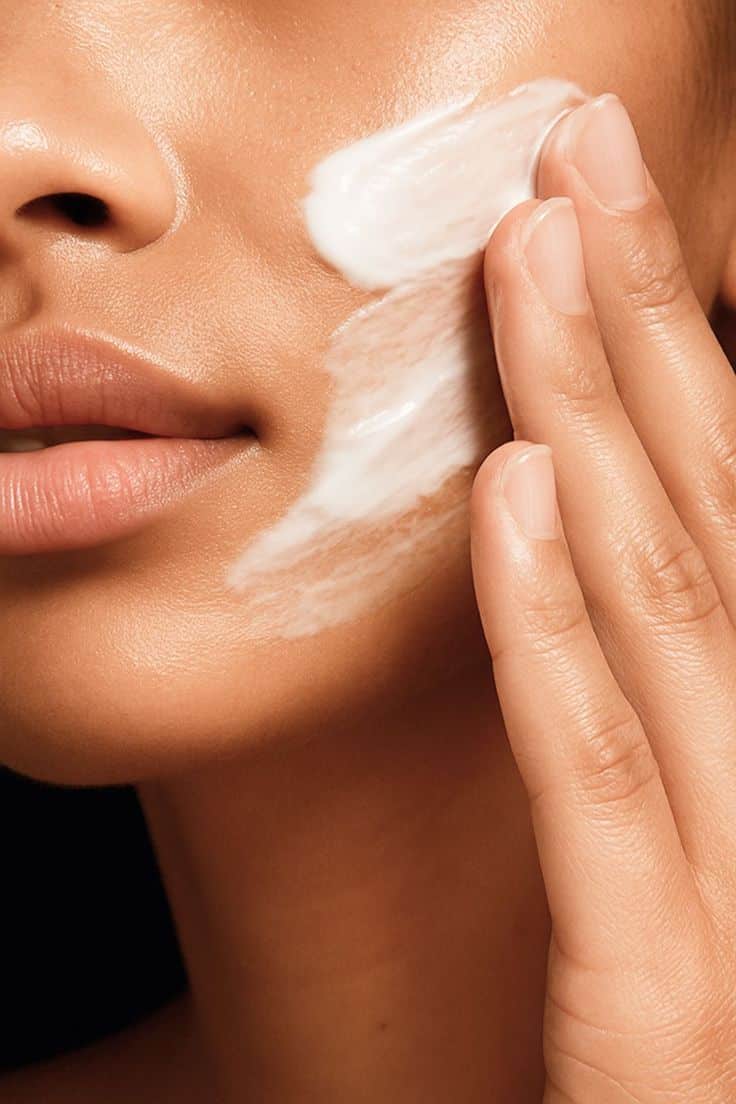 Mão aplicando creme branco na bochecha de pessoa com pele saudável.