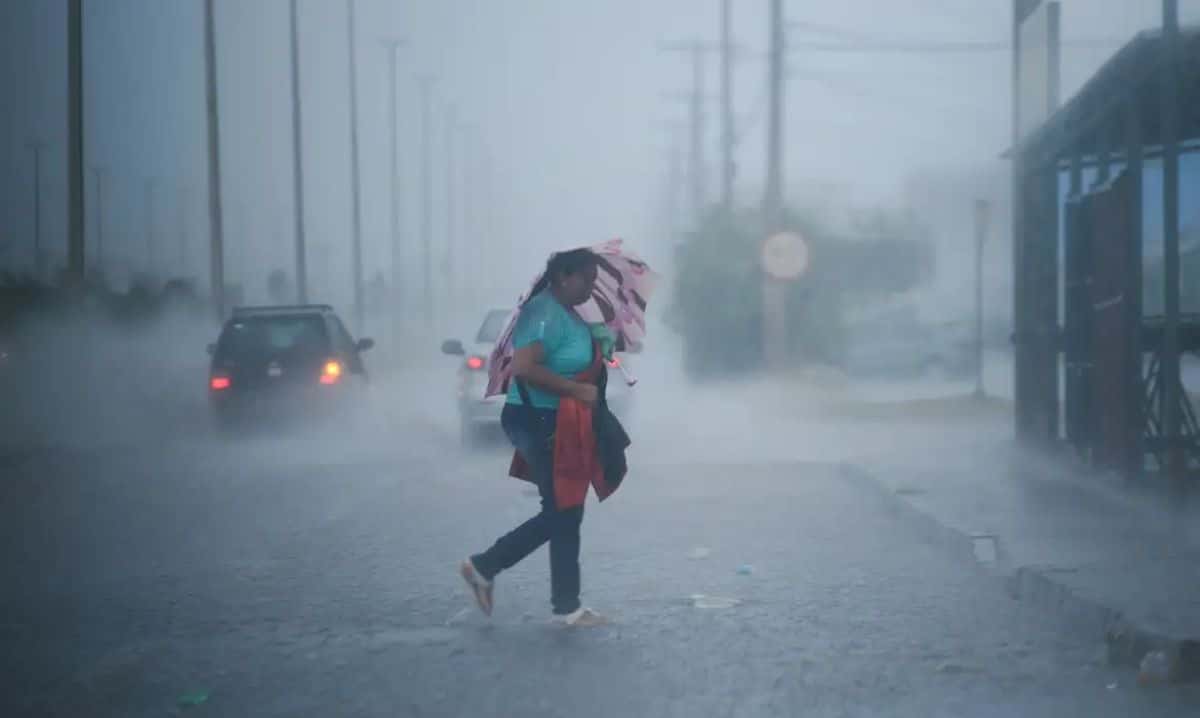Pessoa caminha sob chuva forte em rua alagada durante temporal, com carros ao fundo e baixa visibilidade causada pelo mau tempo.