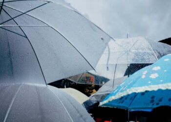 Pessoa com guarda-chuva caminha sob chuva em dia nublado, ilustrando a previsão do tempo para o mês de março no Brasil.