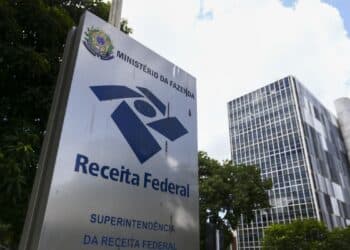 Placa da Receita Federal em frente a prédio moderno rodeado por árvores e céu azul
