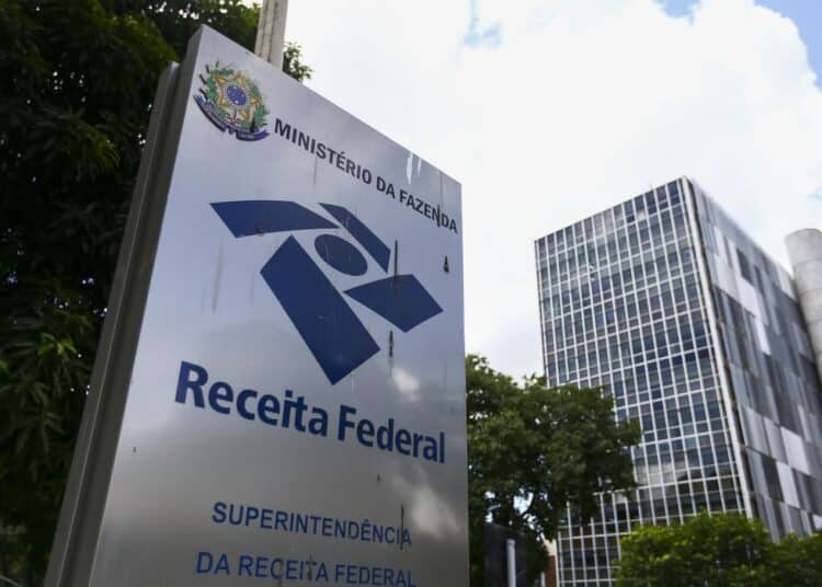 Placa da Receita Federal em frente a prédio moderno rodeado por árvores e céu azul