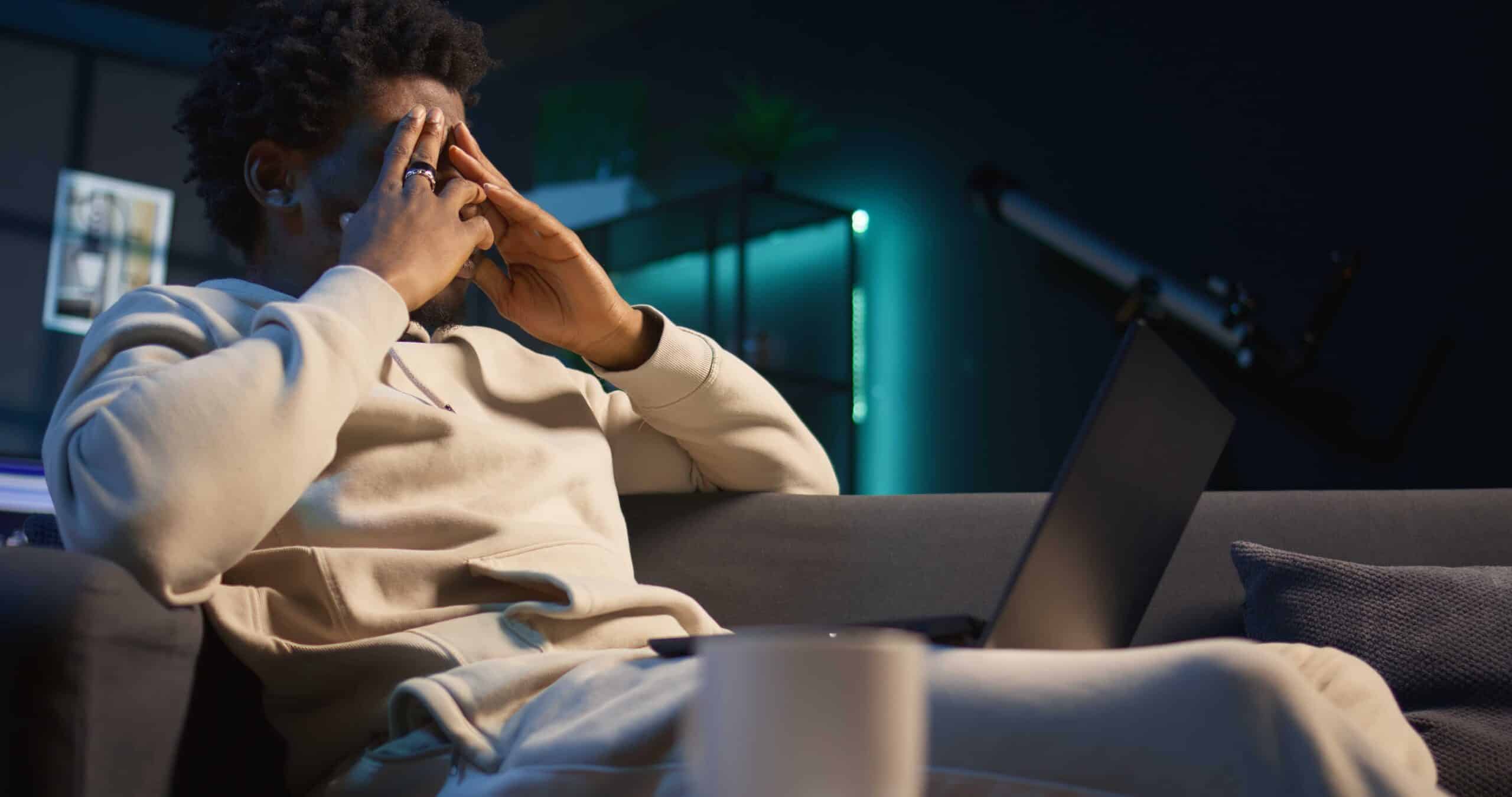 Homem sentado no sofá cobre o rosto com as mãos em frente ao notebook